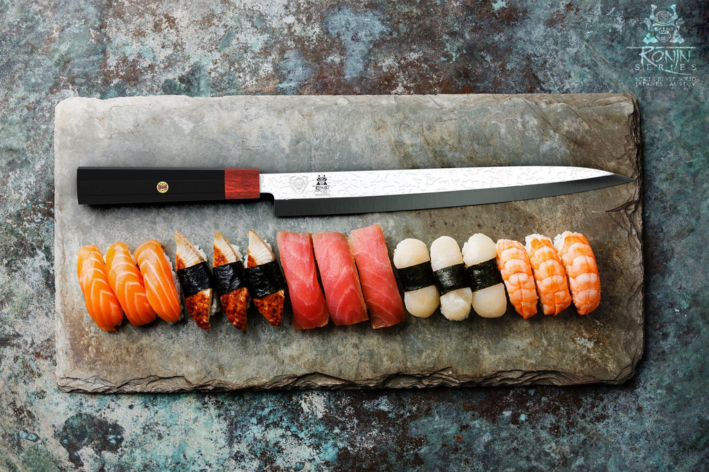Horizontal yanagiba knife on a marble cutting board in front of several sushi pieces of different colors