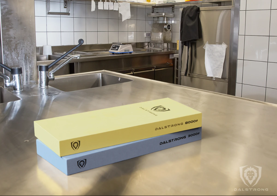 A yellow and blue knife sharpening whetstone on an industrial kitchen surface with an apron hanging in the background