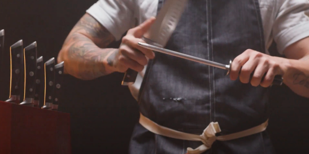 A Chef in a blue apron honing his knife against a sharpening steel with a knife block set next to him