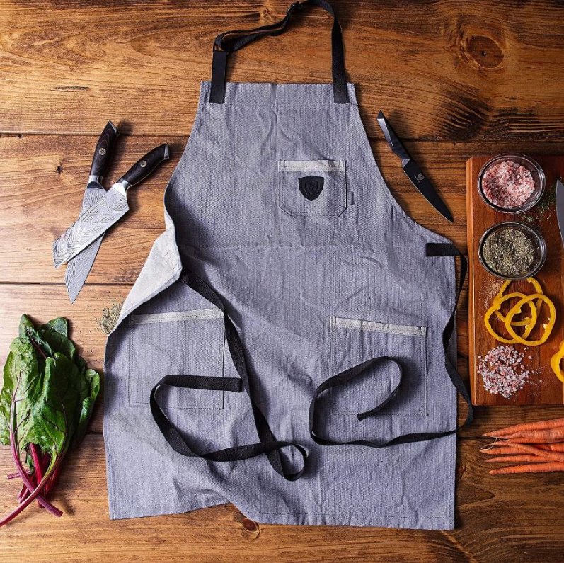 A photo of the American Legend Blue Denim Professional Chef's Kitchen Apron Dalstrong along side with three Dalstrong knives on top of a wooden table.