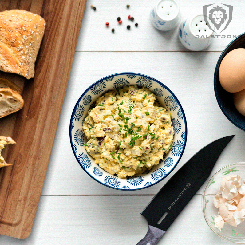 A bowl of egg salad with a Dalstrong Delta Wolf Series knife on the side and slices of bread on a wooden cutting board.