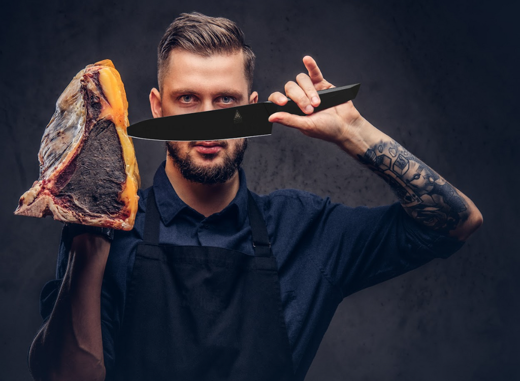 A photo of a man holding Chef's Knife 8" Shadow Black Series | NSF Certified | Dalstrong and a aged meat on the other hand.