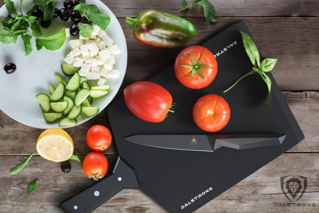 A plate of sliced cucumber and cheese next to a black cutting board with full tomatoes and a black paring knife on it 