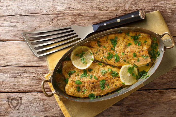 A photo of the Dalstrong Professional Slotted Fish Spatula 7.5" beside of a beautiful cooked fish fillet on top of a wooden table