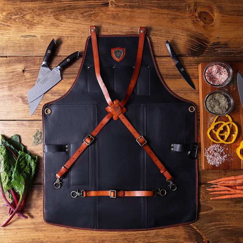 A photo of the The Culinary Commander Top-Grain Leather Professional Chef's Kitchen Apron Dalstrong with three knives beside it in top of a wooden table