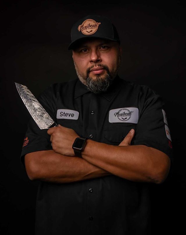 Chef Steve Hernandez in a black tshirt with his arms folded holding a Dalstrong knife