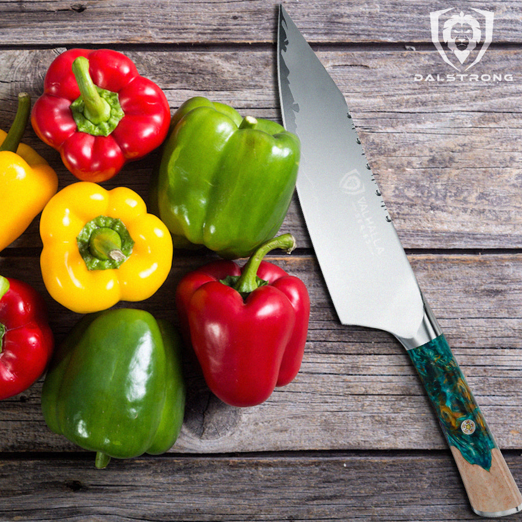 A pile of colourful red green and yellow peppers on a wooden surface next to a sharp kitchen knife