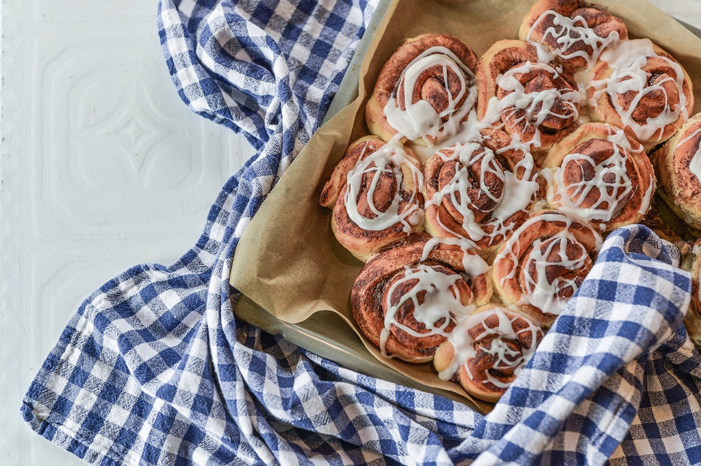 Fresh baked cinnamon buns swirled with cream cheese on top on a baking tray and blue checkered fabric underneath.