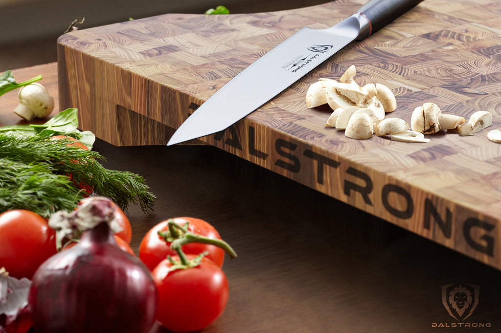 Tomatos and a red onion rest on a table in front of a wooden cutting board that has a chef knife and garlic on top of it