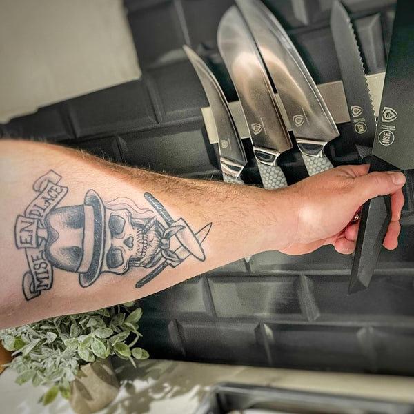 Man reaches for a wall of dalstrong chef knives with a chef knife tattoo on his forearm