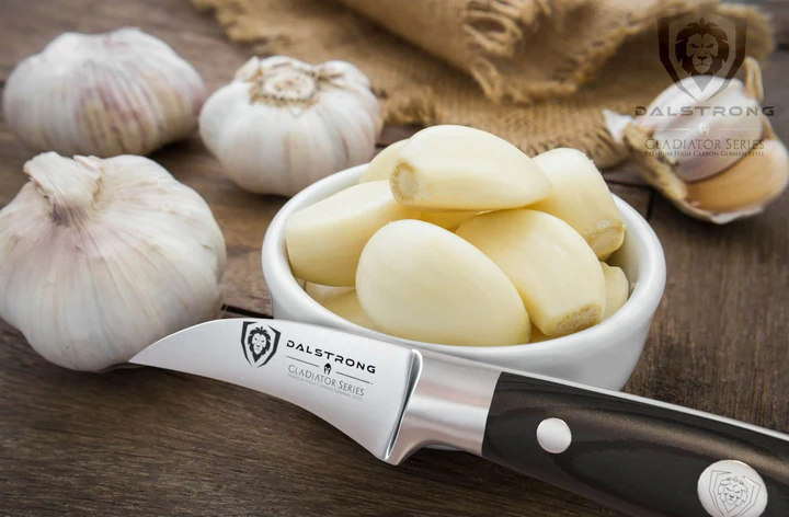 Bunch of garlic cloves inside a ramekin bowl beside of three garlic bulbs and the Bird's Beak Tourne Paring Knife 2.75" Gladiator Series | NSF Certified | Dalstrong on a wooden surface