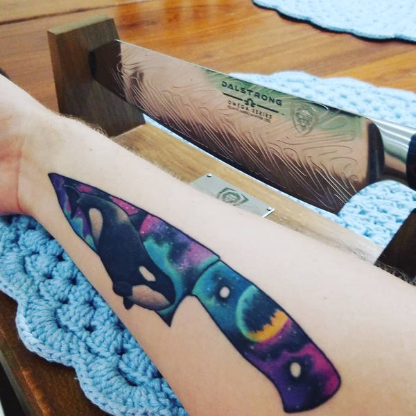 Colorful chef knife tattoo on a forearm next to a real Dalstrong omega series chef knife