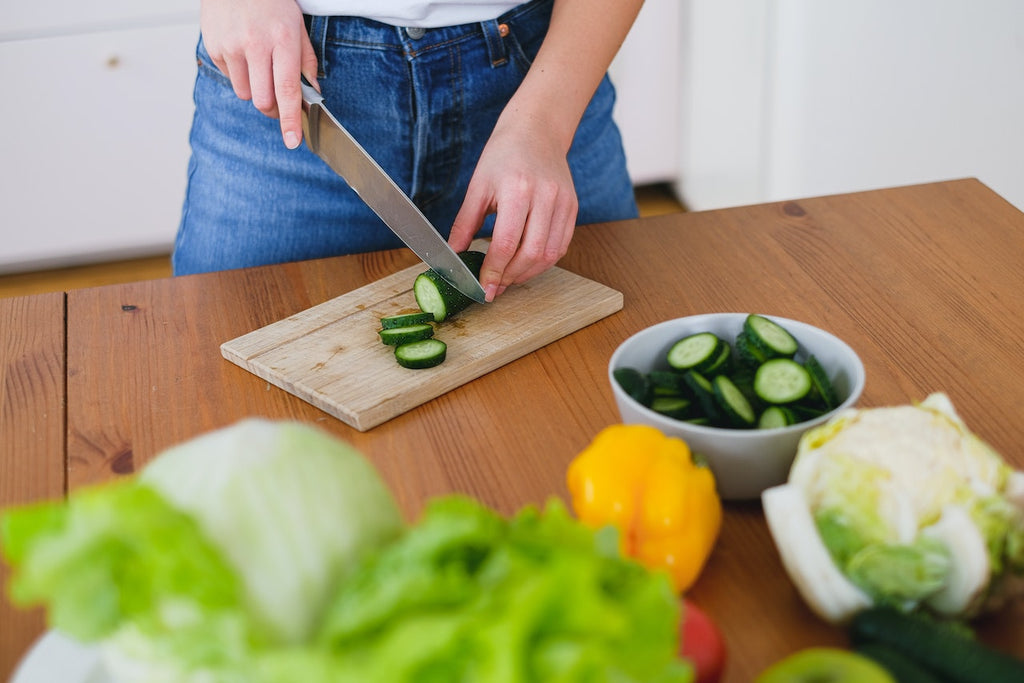 A person slicing a green cucumber on a wooden cutting board with different vegetables on the side
