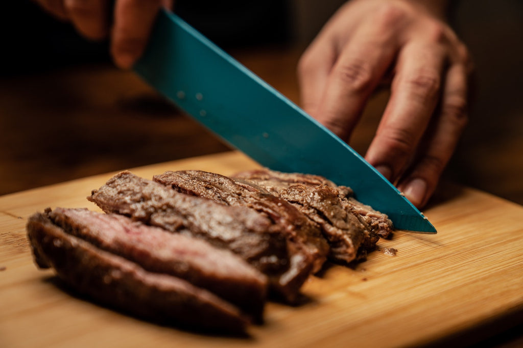 Person slicing meat on a wooden chopping board using a blue ceramic knife