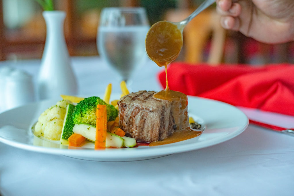 A spoonful of gravy being poured on a steak dish with mixed vegetables.