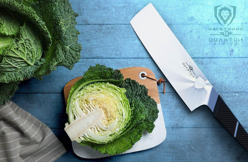 Fresh raw cabbage sliced in half on a wooden cutting board beside a Dalstrong Quantum 1 Series Nakiri knife.