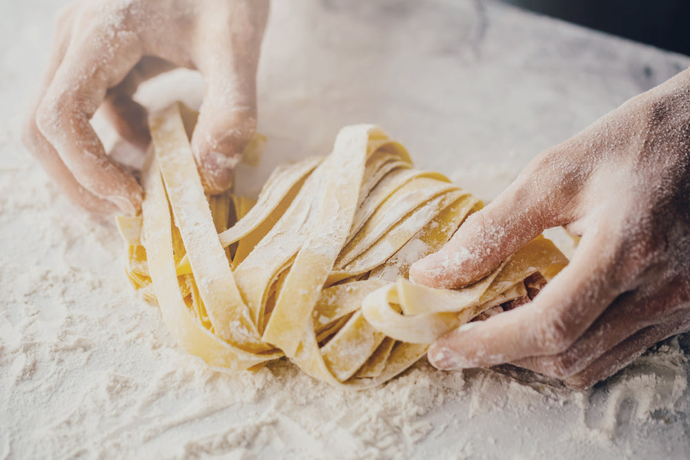 Two hands that are white with flour hold strings of homemade pasta above a table of flour