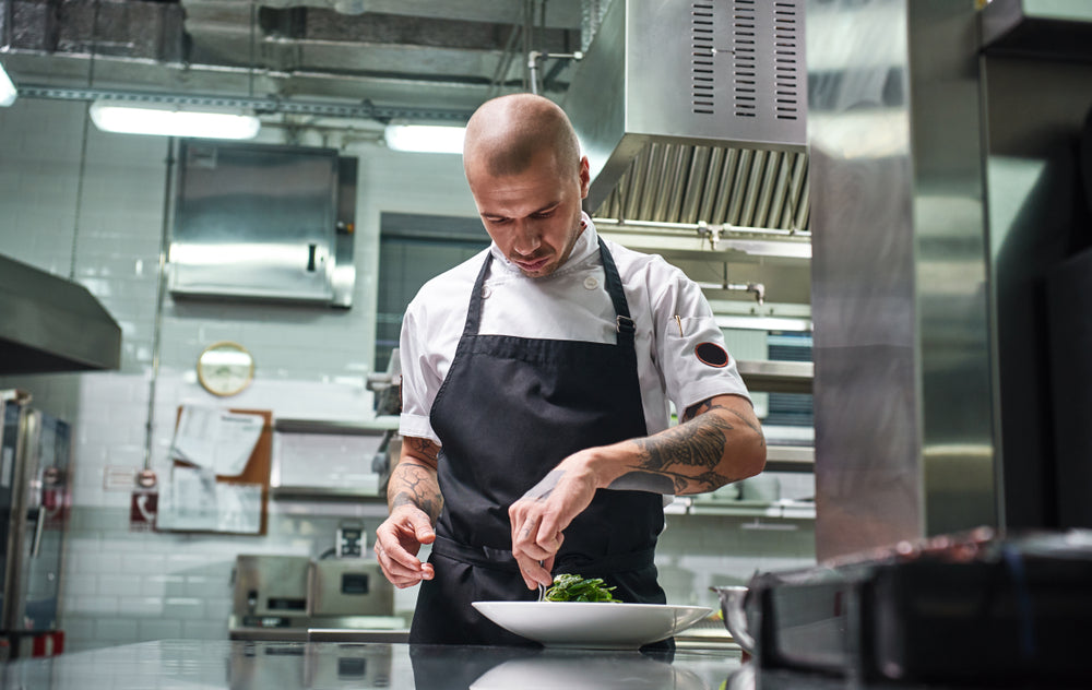 A chef with tattoos prepares a bowl of food in a commercial kitchen