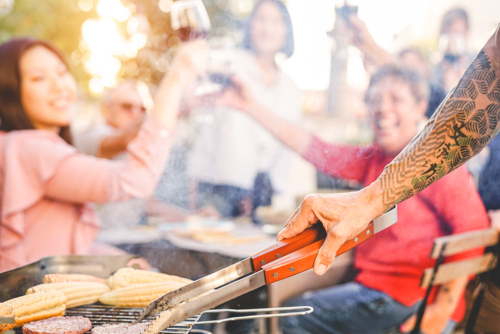 Tattooed arm flipping food on an outdoor grill as people cheer in the background