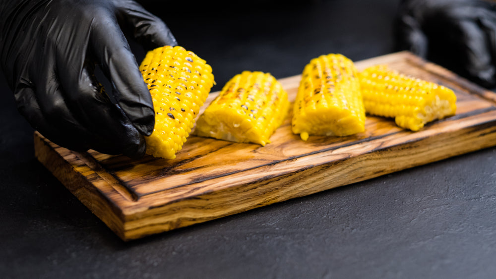 Two hands in black bbq gloves arranging corn on the cob on a small cutting board