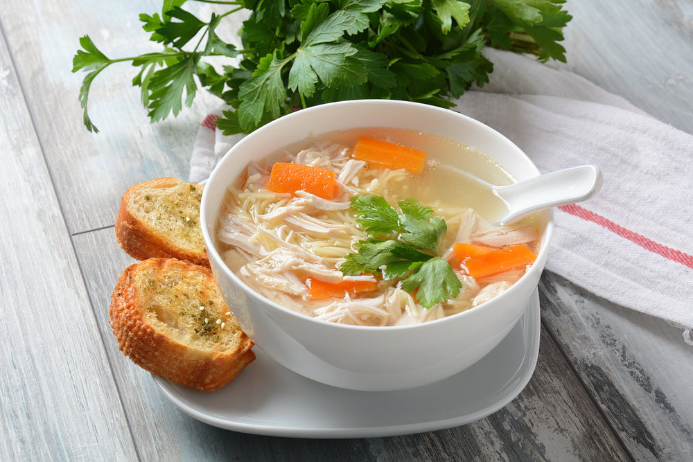 White bowl filled with chicken noodle soup on a white plate with two slices of garlic bread
