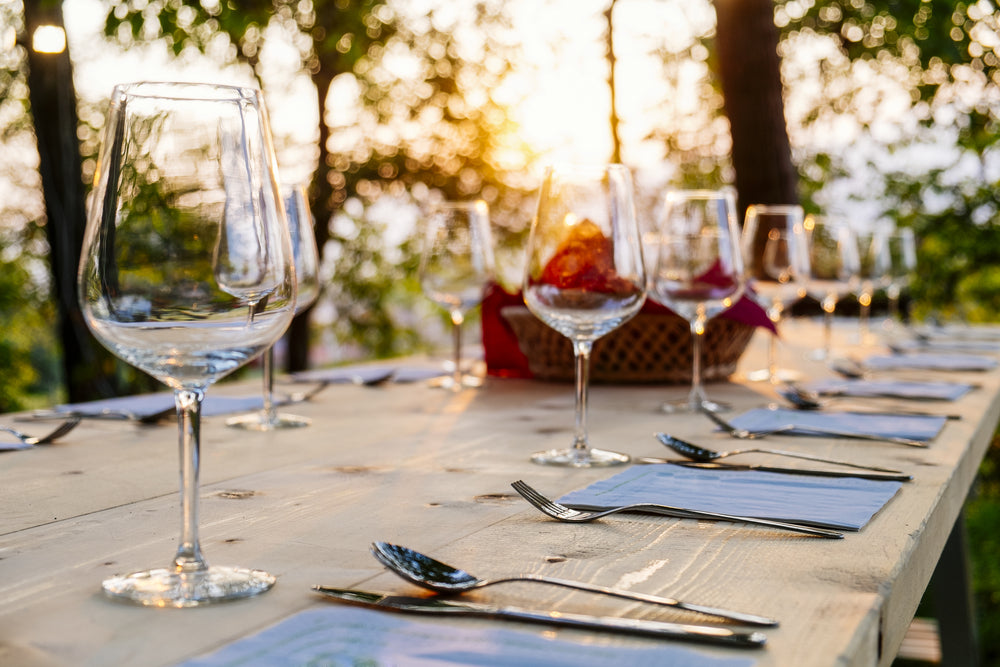 Out door dining table set for dinner with blue place mats, cutlery and wine glasses and a sunset background