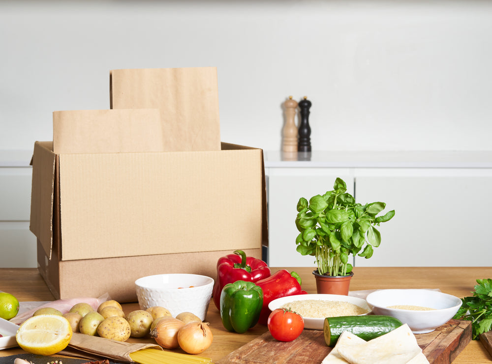 Cardboard box on kitchen counter next to a variety of vegetable ingredients