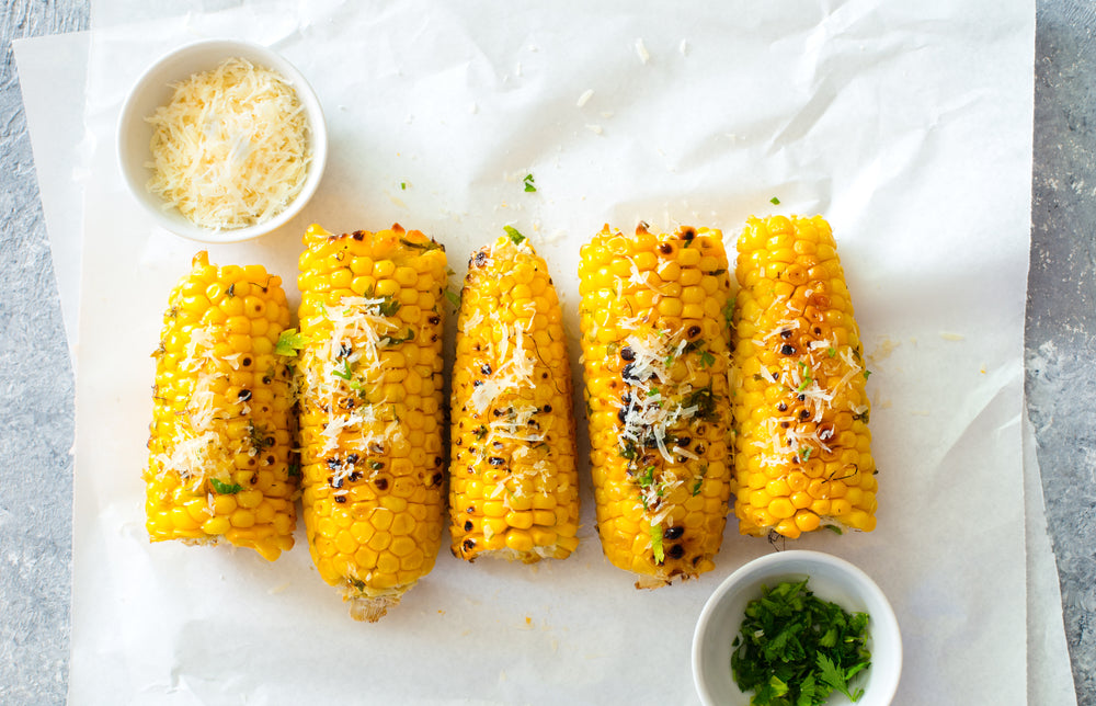 Five pieces of corn on the cob on white parchment paper next to two small bowls of seasoning