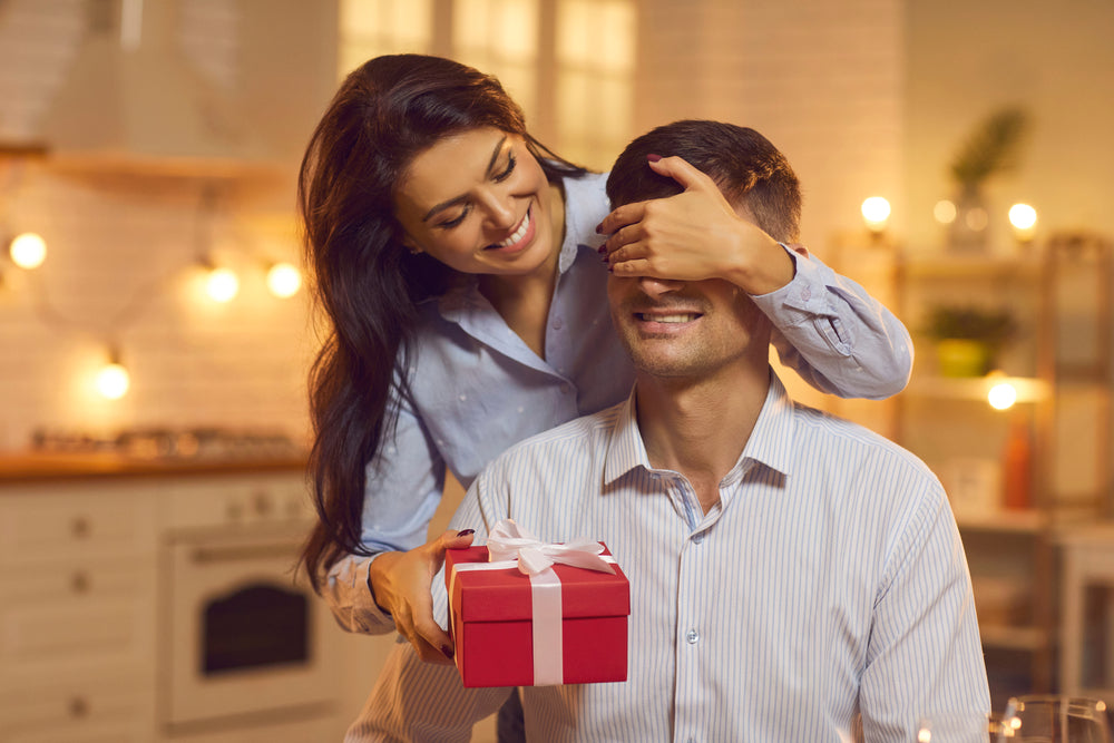 Woman in blue shirt standing in kitchen covers boyfriend's eyes to surprise him with red gift box