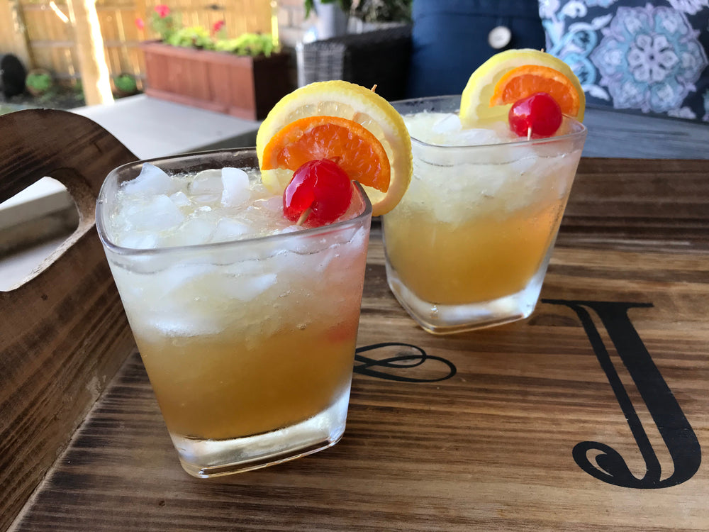 Two John Collins cocktails on a wooden cutting board with an outdoor background