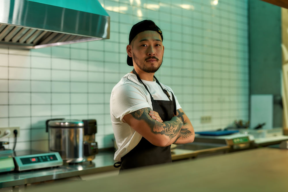 A chef with tattoos stands facing the camera with his arms folded