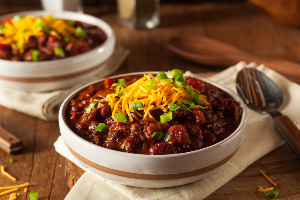 Two white bowls filled with chili and cheese next to a white napkin and a spoon on a wooden table