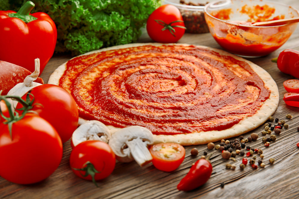 Uncooked pizza dough with tomato sauce spread across it surrounded by fresh tomatoes and diced mushrooms