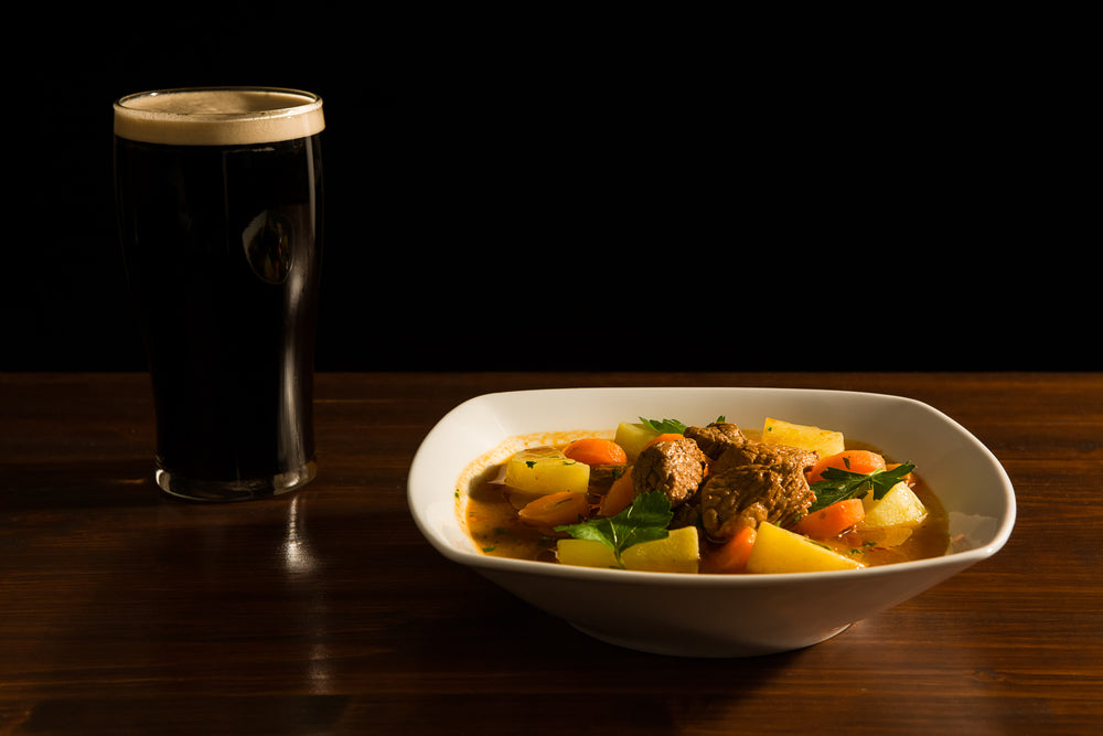 Pint glass of dark beer with white head next to a bowl of traditional Irish beef stew