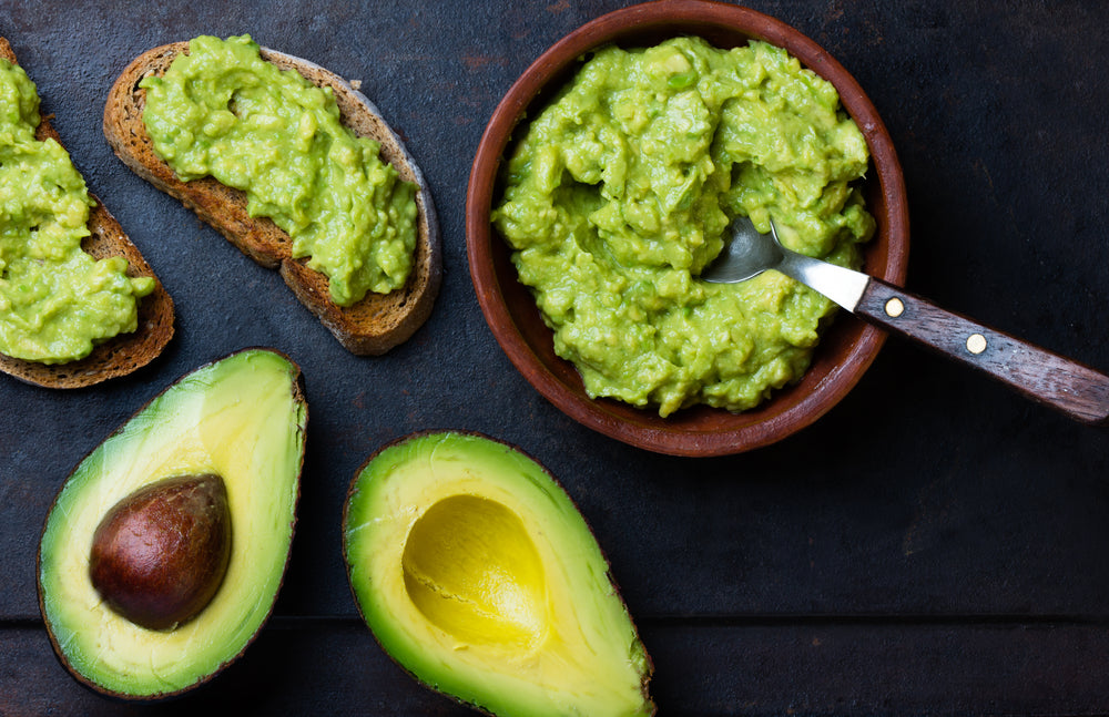 A bowl of guacamole next to chopped avocados and bread with guacamole topping