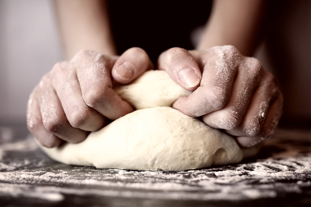 Two hands work on a mound of pizza dough with flour spread across the table