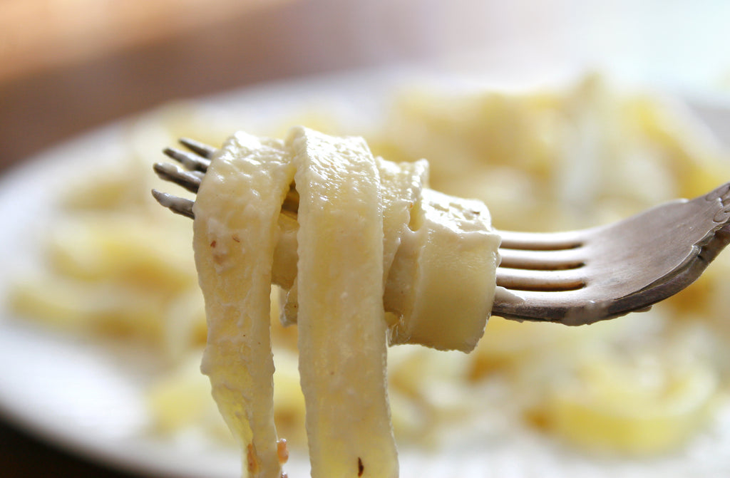 Pasta covered in alfredo sauce wrapped around a silver fork with a blurred plate of past a in the background