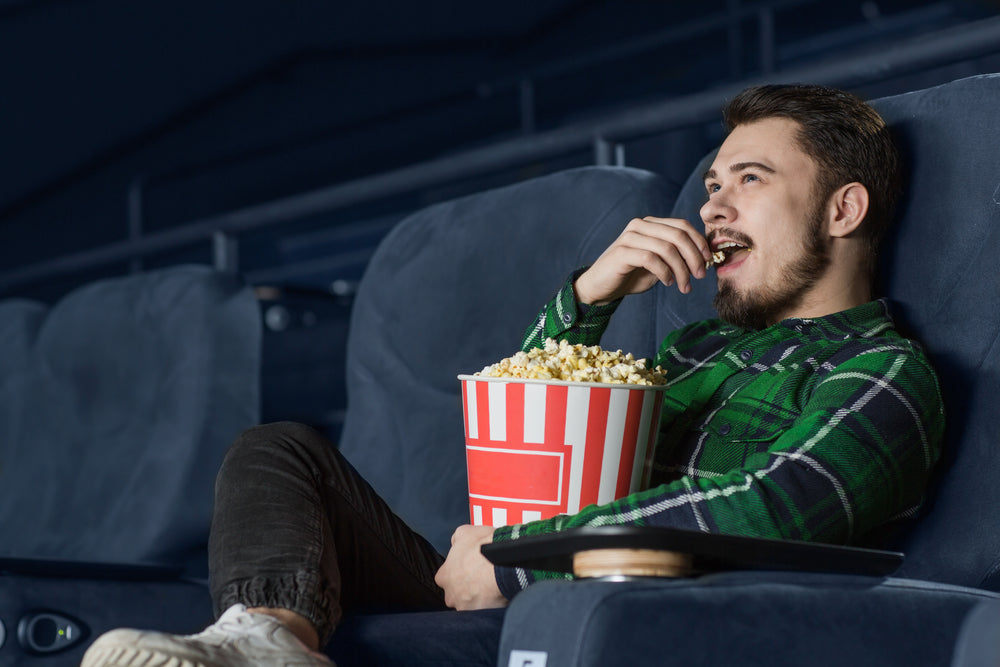 Bearded man wearing green checkered shirt smiles as he eats popcorn in a movie theatre