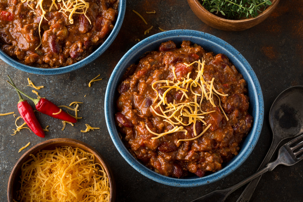 Bowls of chili with cheese on top next to a bowl of cheese and with peppers on the table