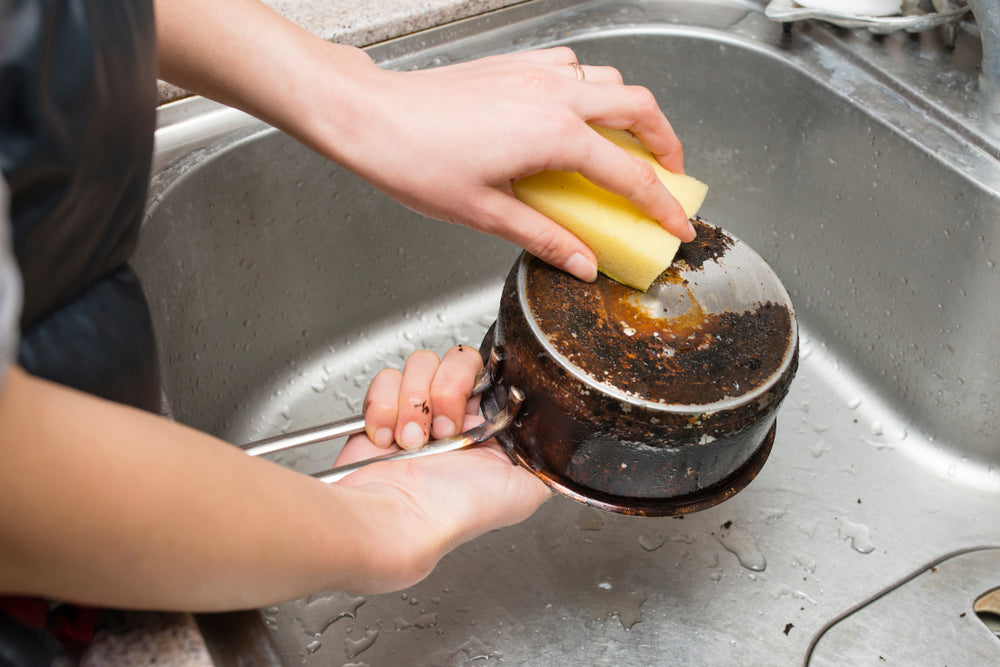 Hands using a yellow sponge over a sink to clean the bottom of a burned saucepan