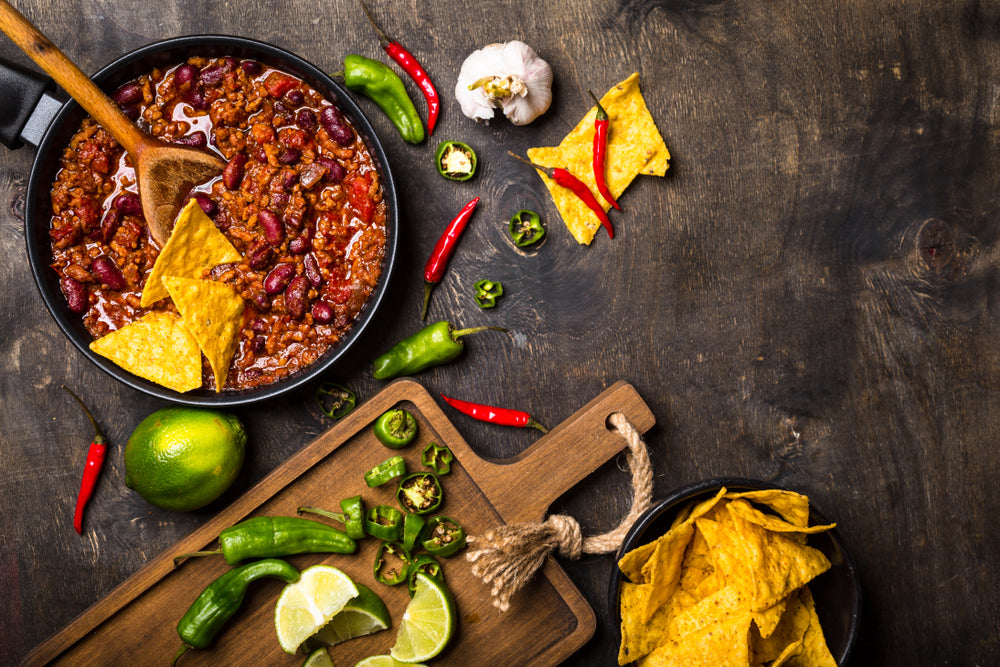 A large bowl of chili with a wooden spoon sticking out of it next to a cutting board with chopped limes and vegetables 