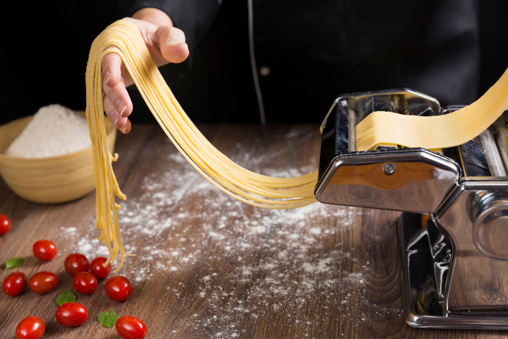 Chef making spaghetti noodles with pasta machine on kitchen table with some ingredients around.