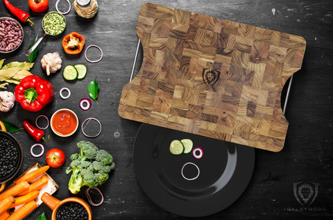 Dalstrong Teak Cutting board on wooden surface surrounded by vegetables 