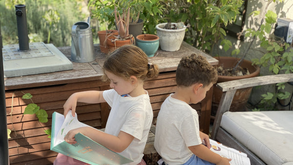 Two kids are reading books on a terrace outside. Earth month, kids activities, outdoor kids activities