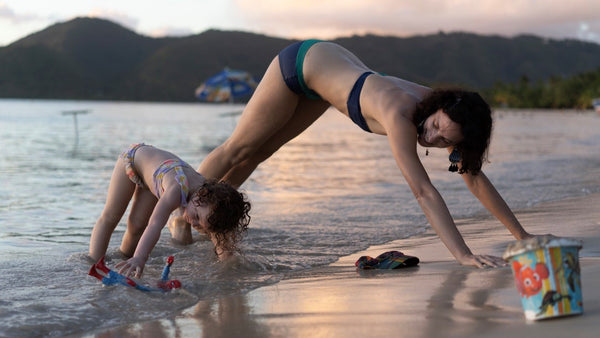 A Mom and her toddler daughter are doing yoga at the beach. Family, travel tips.