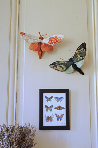two finished moths hanging from nails on the wall, just above a print of butterflies and a large vase of lavender
