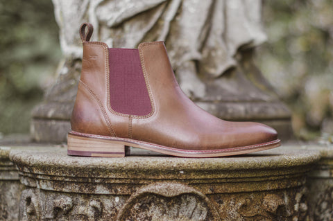 A brown Chelsea boot with burgundy elastic panel, stood on a stone plinth in a graveyard.