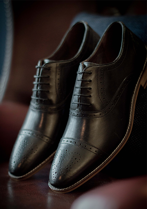 A Pair of London Brogue Artheur Oxford shoes in black with black laces. The shoes are leaning uprightagainst the back of a red leather chair so that you can see the full detailing of the broguing on the toe-caps and side of the shoes.