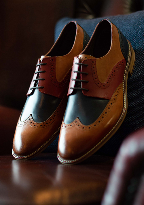 A pair of London Brogue Tommy Derby Shoes in Tan blue and red leather leaning upright with their soles against the back of a red leather chair so you can see the full brogue detail on the toes of the shoes