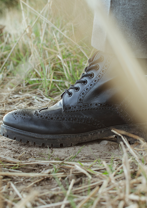 A close up photo of a right London Brogue Billy Boot in Black under the cuff of a tweed trouser leg. Stood in a field with strands of grass in the foreground 